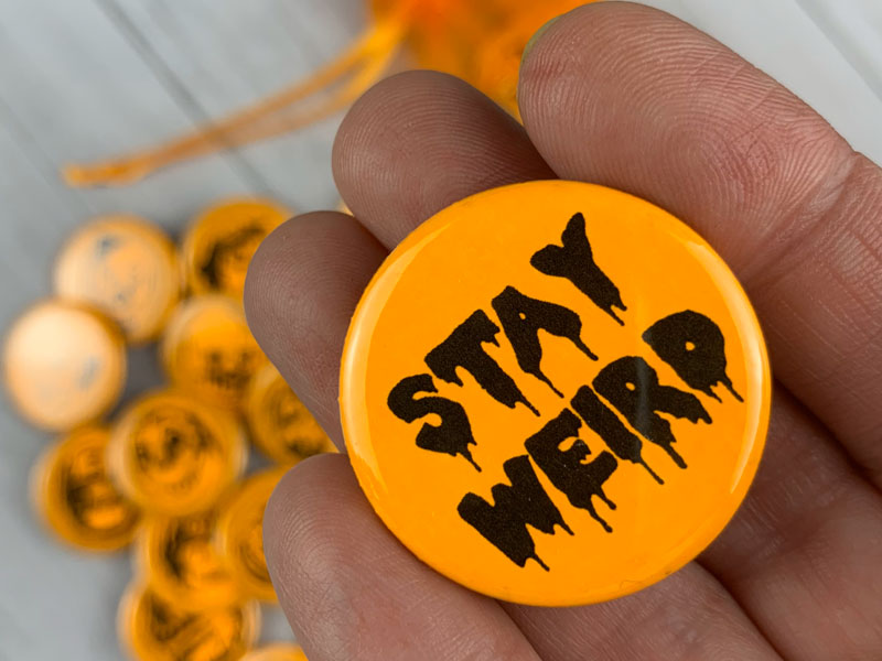 Round neon orange pin with phrase Stay Weird in a drippy horror font.